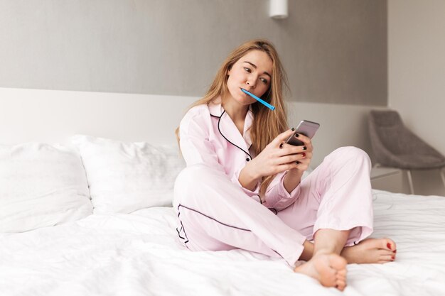 Portrait of young lady sitting on bed with toothbrush while using cellphone at home