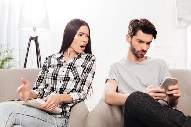 Portrait of young lady sitting in chair with book in hand and amazedly looking in cellphone of near sitting man at home isolated