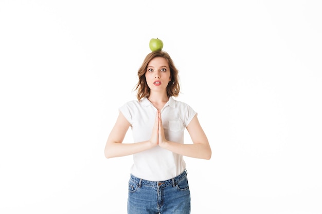 Free photo portrait of young lady standing with green apple on head and amazedly looking in camera on white background isolated