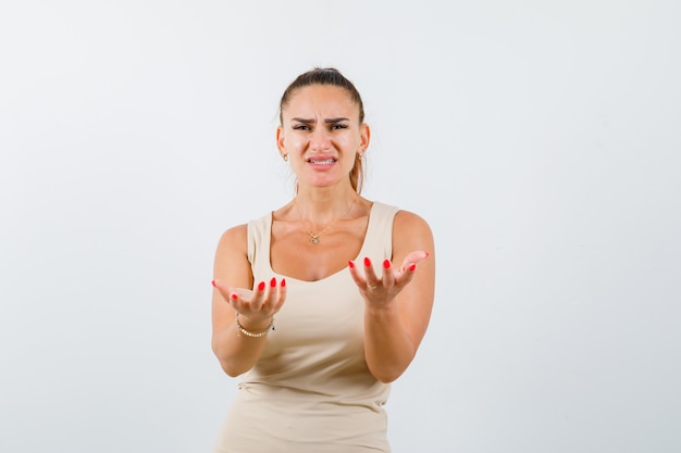 Portrait of young lady stretching hands towards camera in beige tank top and looking mournful front view