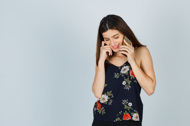 Portrait of young lady talking on mobile phone in blouse and looking ashamed front view