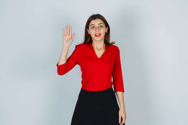 Portrait of young lady waving hand for greeting in red blouse, skirt and looking happy