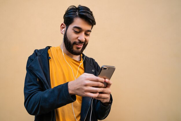 黄色の空間に対してイヤホンで彼の携帯電話を使用して若いラテン男の肖像画。コミュニケーションの概念。