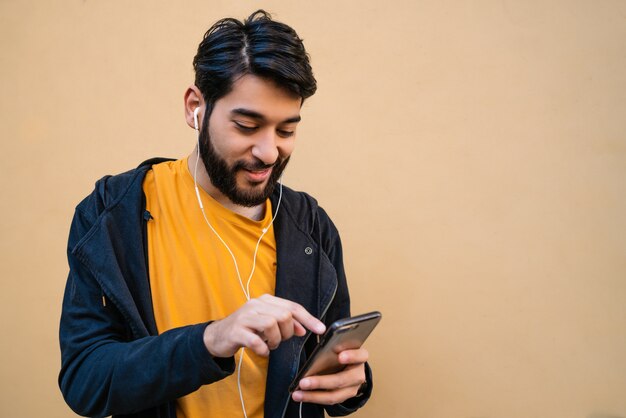 Portrait of young latin man using his mobile phone with earphones against yellow space. Communication concept.