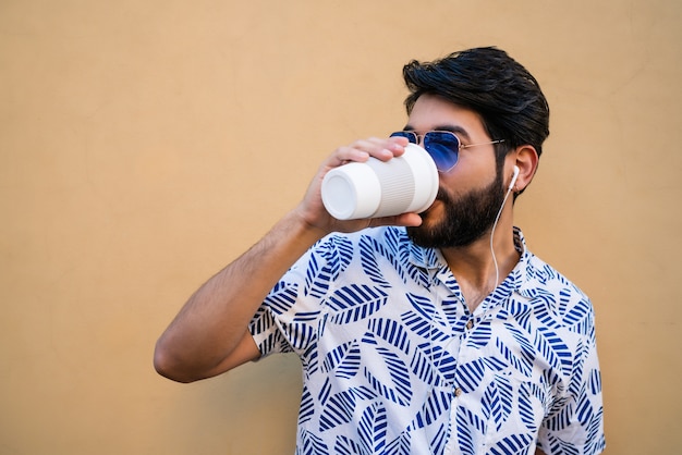 Portrait of young latin man wearing summer clothes, drinking a cup of coffee and listening to music with earphones against yellow space.