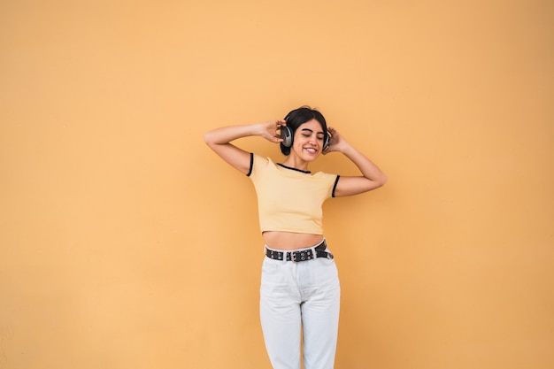 黄色の空間に対してヘッドフォンで音楽を聴く若いラテン女性の肖像画。都市のコンセプト。
