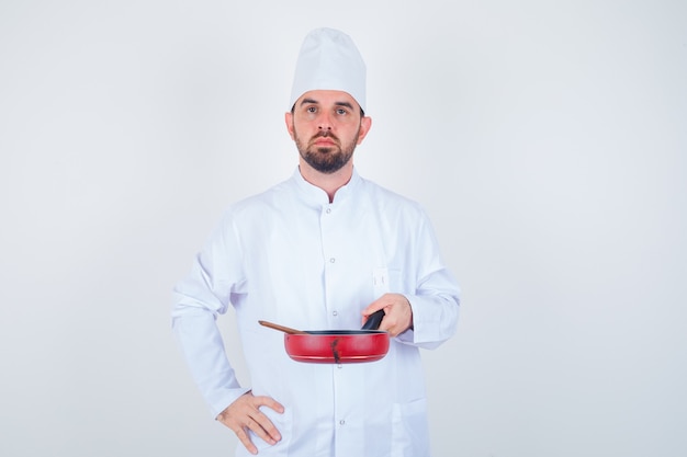 Portrait of young male chef holding frying pan with wooden spoon in white uniform and looking serious front view