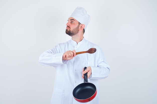 Portrait of young male chef threatening with frying pan and wooden spoon in white uniform and looking nervous front view