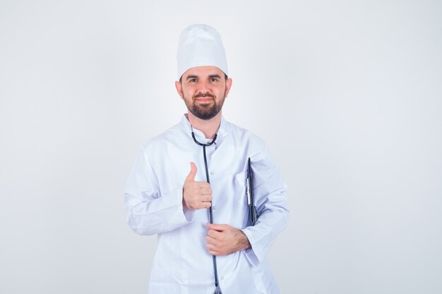 Портрет молодого мужчины-врача, держащего буфер обмена, показывающего большой палец вверх в белой форме и веселого вида спереди