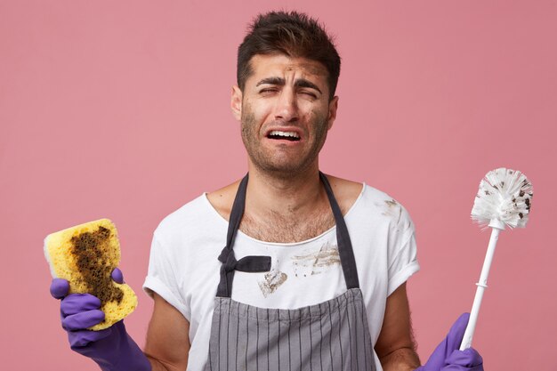 Portrait of young male doing chores having sorrowful expression not wanting to clean everything. Male wearing apron holding sponge with brush having tired and frustrated look