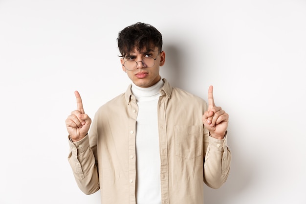 Portrait of young male model in glasses looking suspicious and frowning, pointing fingers up with disbelief, standing on white background.