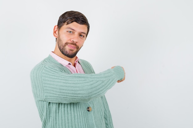 Portrait of young male pointing back in shirt, cardigan and looking cheery front view