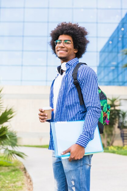 キャンパスに対して使い捨てのコーヒーカップと本を手で保持している若い男子学生の肖像画