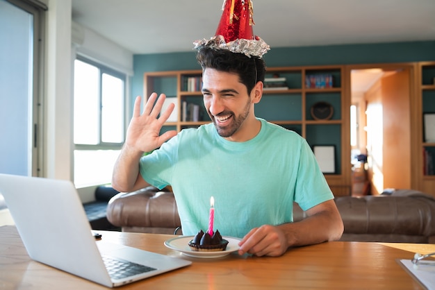 Portrait of young man celebrating birthday on a video call from home with laptop and a cake. New normal lifestyle concept.