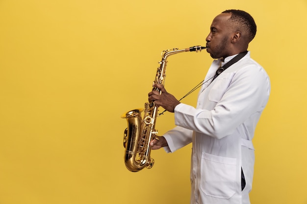 Portrait of young man doctor with saxophone isolated on yellow