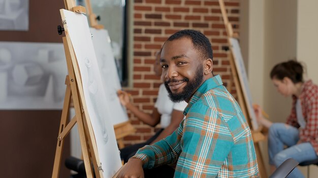 鉛筆、帆布、イーゼルを使用して芸術的なスキルを開発するアートクラスでインスピレーションオブジェクトを描く若い男の肖像画。開発のためのスケッチを描くことを学ぶための余暇活動。ハンドヘルドショット