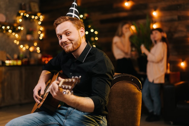 Portrait of young man in the festive hat playing on the guitar against the background of talking friends. christmas tree with garland and wall with festive illumination in background.