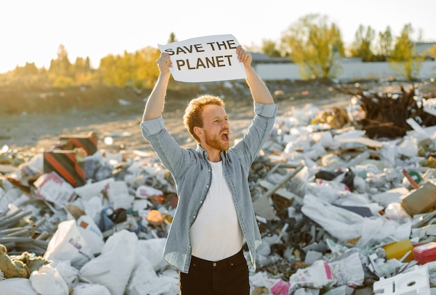 Portrait of young man fighting for nature holding save mother earth sign at dump. protesting against nature pollution waving hands calling on to save planet.