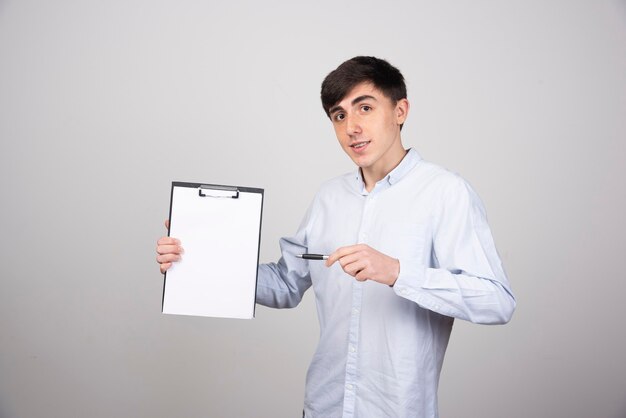 Portrait of young man holding empty clipboard on gray wall.