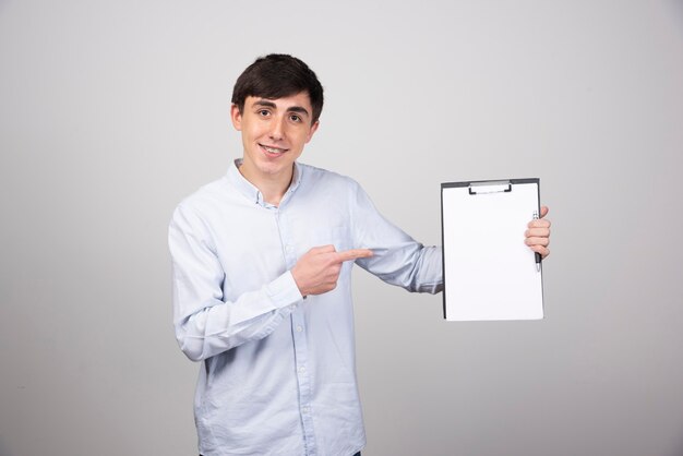 Portrait of young man holding empty clipboard on gray wall.