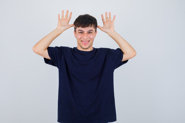 Портрет молодого человека, держащего руки возле головы как уши, высунувшего язык в черной футболке и удивленного вида спереди