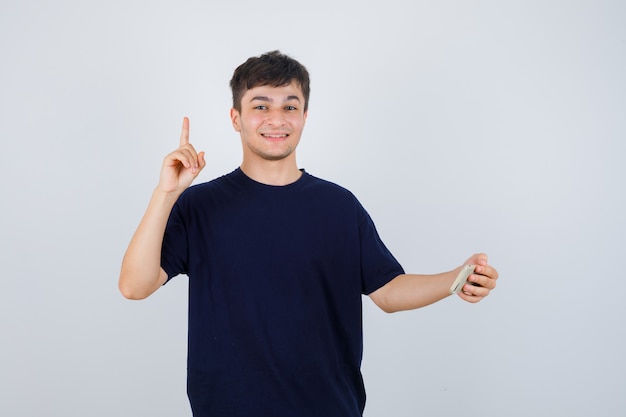 Портрет молодого человека, держащего мобильный телефон, указывая вверх в черной футболке и уверенно выглядящего, вид спереди