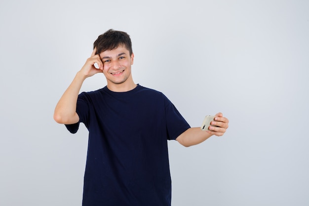 Portrait of young man holding mobile phone, standing in thinking pose in black t-shirt and looking cheerful front view