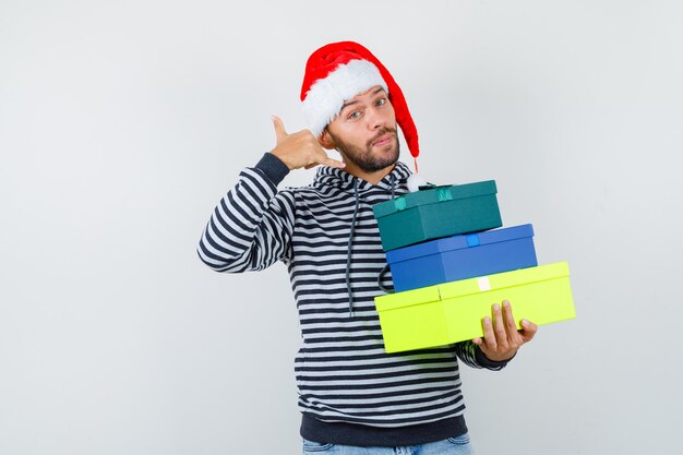 Portrait of young man holding present boxes, showing phone gesture in hoodie, Santa hat and looking confident 