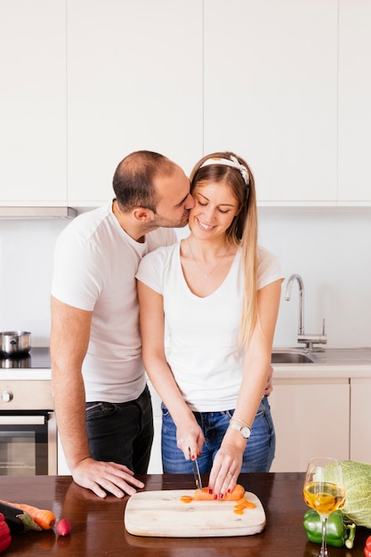 Free photo portrait of a young man kissing his girlfriend cutting the carrot with knife on wooden table in the kitchen