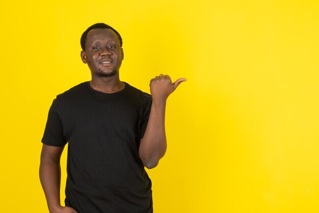 Portrait of a young man model standing and pointing away with thumb against yellow wall 