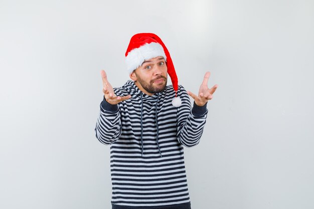 Portrait of young man opening arms for hug in hoodie, Santa Claus hat and looking sincere 