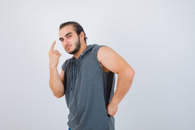 Portrait of young man pointing up while holding hand on waist in sleeveless hoodie and looking sensible front view