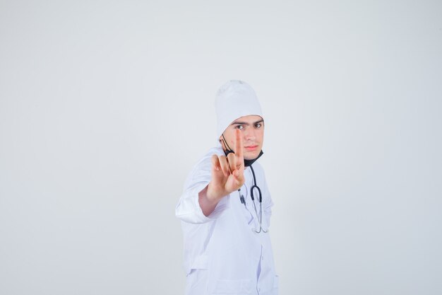 Portrait of young man pointing up in white uniform, mask and looking confident front view