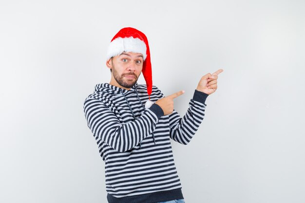 Portrait of young man pointing at upper right corner in hoodie, Santa hat and looking puzzled 
