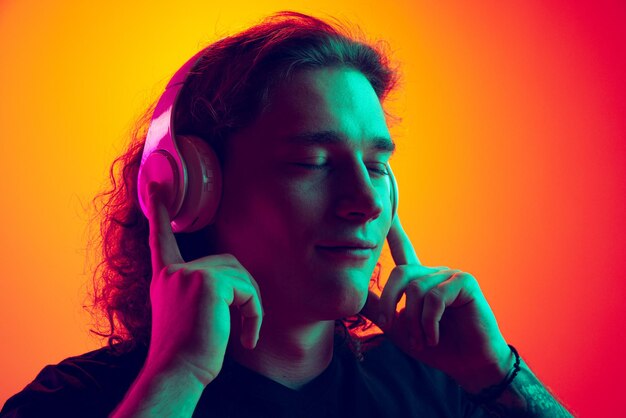 Portrait of young man posing listening to music in headphones isolated over gradient red orange background in neon light