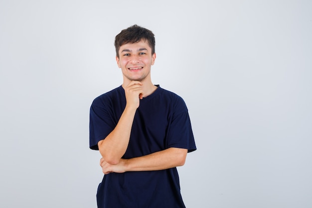 Portrait of young man propping chin on hand in black t-shirt and looking cheerful front view