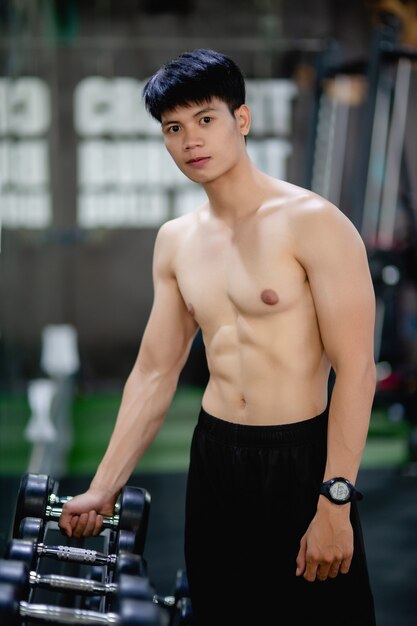 Portrait young man shirtless taking dumbbell from rack for exercise at fitness gym,, 