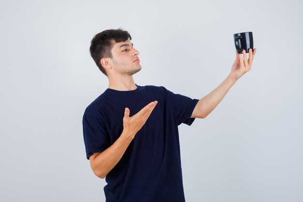 Portrait of young man showing cup of tea in black t-shirt and looking pensive front view