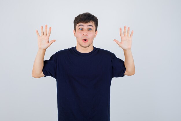 Portrait of young man showing surrender gesture in black t-shirt and looking scared front view