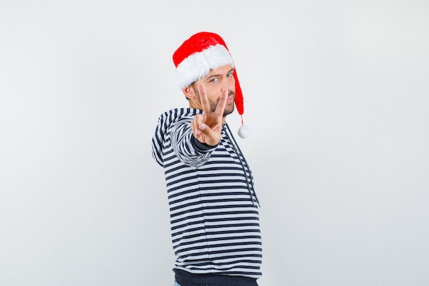 Portrait of young man showing victory gesture in hoodie, Santa hat and looking confident