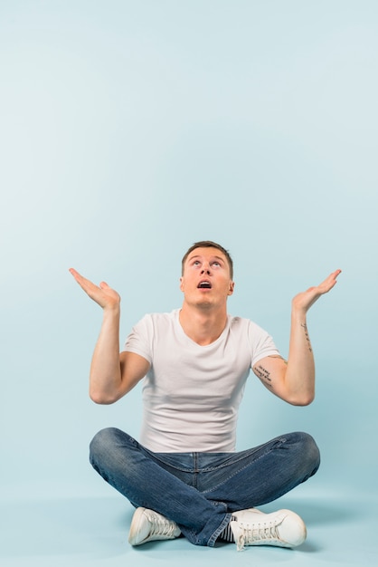 Portrait of a young man sitting down with crossed legs looking up shrugging