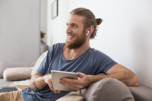 イヤホンとタブレットを手に灰色のソファに座って、家で幸せに脇を見て若い男の肖像画