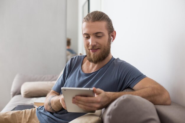 Portrait of young man sitting on gray sofa with earphones and the tablet in hands at home