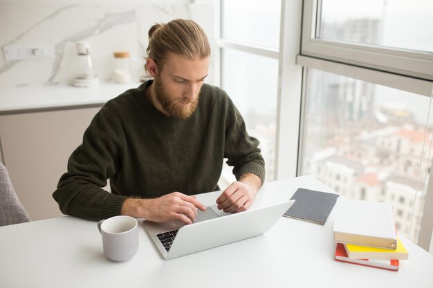 Portrait of young man sitting at the table with cup and books while working on laptop at home