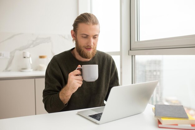 手にカップと自宅でラップトップとテーブルに座っている若い男の肖像画