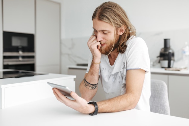 Portrait of young man sitting with digital tablet in hand in kitchen at home