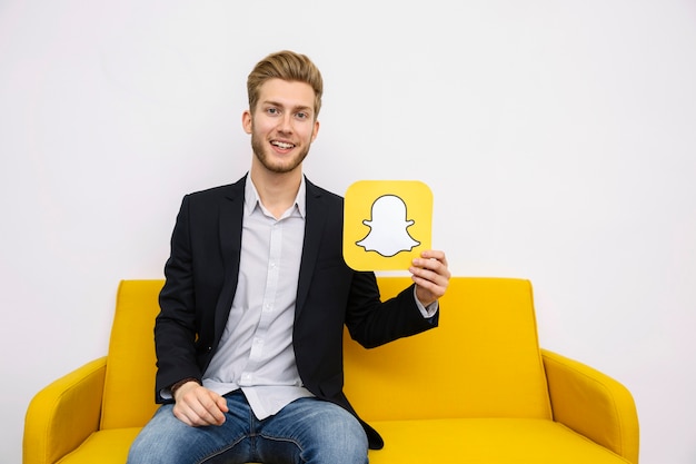 Portrait of young man sitting on yellow sofa holding snapchat icon