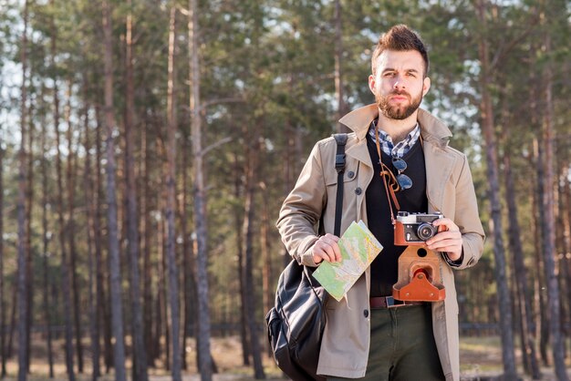 Portrait of a young man standing in the forest holding map and vintage camera