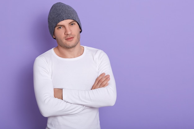 Free photo portrait of young man standing with hands folded on lilac, attarctive male wearing white long sleevw shirt and gray cap. copy space for advertisment or promotion text.