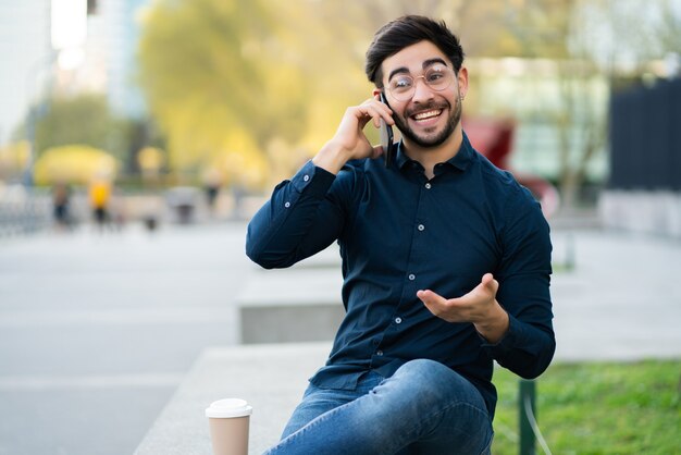 Portrait of young man talking on the phone while sitting on bench outdoors. Urban concept.